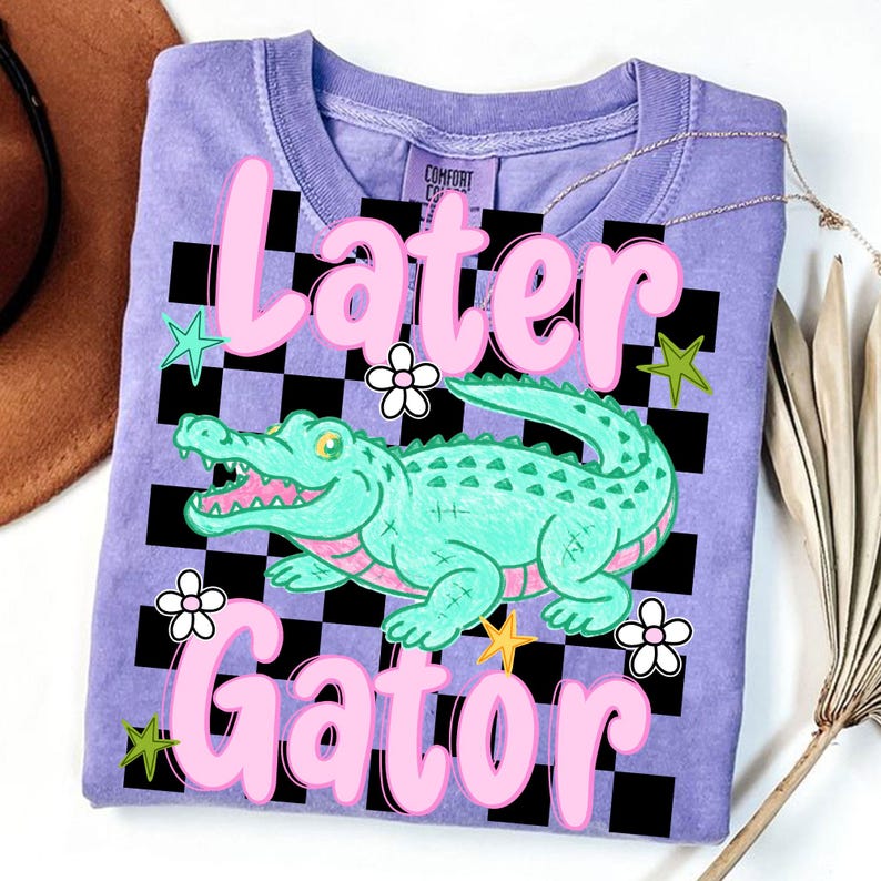 May include: A lilac-colored t-shirt with the words "Later Gator" in pink, featuring a cartoon alligator. The design includes a black and white checkered background, green stars, and white flowers.