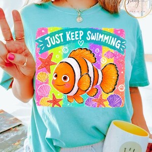 K&ouml;nnte beinhalten: Hellblaues T-Shirt mit einem farbenfrohen Aufdruck eines Cartoon-Clownfischs und dem Text "JUST KEEP SWIMMING". Das Design beinhaltet Seesterne und Muscheln. Das Shirt ist ein l&auml;ssiger Stil.