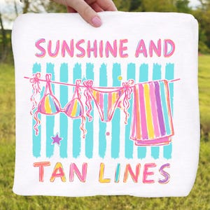 Peut inclure: Serviette de plage blanche avec une illustration color&eacute;e d'un bikini, d'un bas de bikini et d'une serviette ray&eacute;e suspendus &agrave; une corde &agrave; linge. Le texte "SUNSHINE AND TAN LINES" est &eacute;crit en rose et jaune.