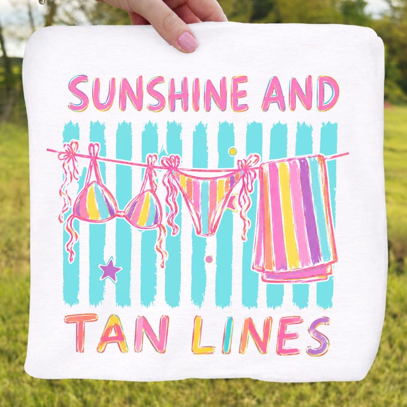 Peut inclure: Serviette de plage blanche avec une illustration color&eacute;e d'un bikini, d'un bas de bikini et d'une serviette ray&eacute;e suspendus &agrave; une corde &agrave; linge. Le texte "SUNSHINE AND TAN LINES" est &eacute;crit en rose et jaune.