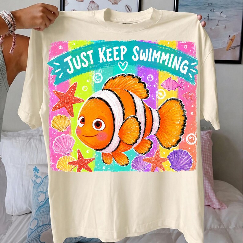 K&ouml;nnte beinhalten: Cremefarbenes T-Shirt mit einer bunten Grafik eines orange-wei&szlig; gestreiften Clownfischs und dem Text "Just Keep Swimming". Das Design umfasst Seesterne, Muscheln und andere Fische auf gestreiftem Hintergrund.