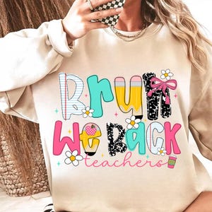 May include: A beige sweatshirt with a colorful graphic that reads "BRU WE BACK teachers". The text features a mix of fonts and designs, including a pencil, a notebook, and a pink bow. Daisies and a coffee cup are also included.