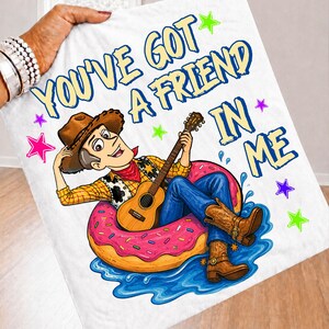 May include: A white blanket featuring a cartoon cowboy wearing a brown hat, yellow shirt, and blue jeans, sitting in a pink donut floatie on blue water. The cowboy plays a brown acoustic guitar. The text reads YOU'VE GOT A FRIEND IN ME.