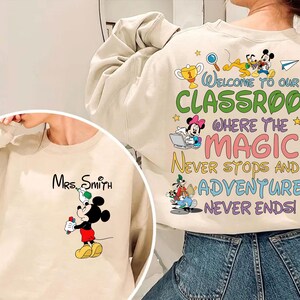 May include: Beige sweatshirt featuring Mickey Mouse holding a marker, with "Mrs. Smith" printed above. The back displays colorful text: "Welcome to our classroom where the magic never stops and the adventure never ends!" with Disney characters.