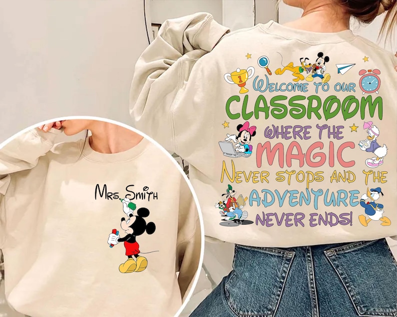 May include: Beige sweatshirt featuring Mickey Mouse holding a marker, with "Mrs. Smith" printed above. The back displays colorful text: "Welcome to our classroom where the magic never stops and the adventure never ends!" with Disney characters.