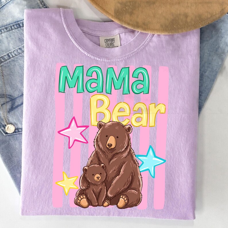 Pu&ograve; includere: T-shirt lilla con le parole "MAMA Bear" in verde acqua e giallo, con un'illustrazione di un orso bruno e un cucciolo. Strisce verticali rosa e stelle circondano il testo e l'illustrazione dell'orso.