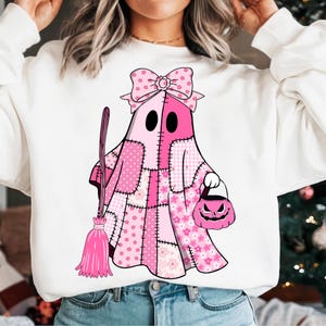 May include: White sweatshirt featuring a pink ghost character with a patchwork design. The ghost holds a pink broom and a jack-o'-lantern bag. The ghost has a bow on its head, perfect for Halloween.
