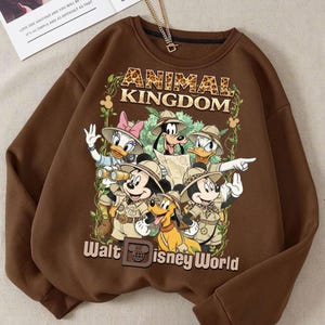 May include: Brown sweatshirt featuring a graphic of Disney characters in safari outfits with the text "ANIMAL KINGDOM" and "Walt Disney World". The design includes Mickey Mouse, Minnie Mouse, Donald Duck, Daisy Duck, Goofy, and Pluto.