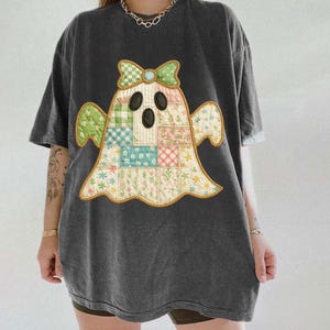 May include: Dark gray t-shirt featuring a patchwork ghost design. The ghost has a bow and is made of floral and checkered patterns in pastel colors. The shirt has short sleeves and a relaxed fit, suitable for casual wear.