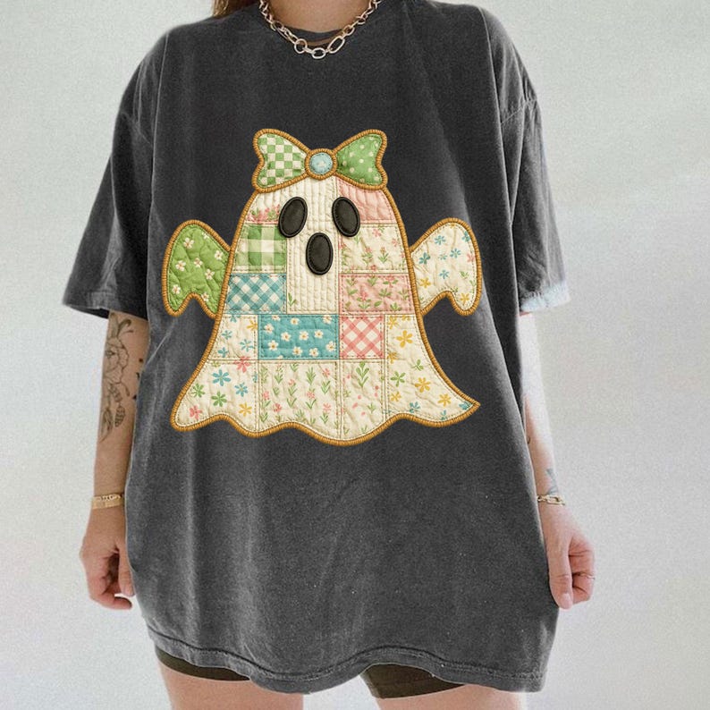 May include: Dark gray t-shirt featuring a patchwork ghost design. The ghost has a bow and is made of floral and checkered patterns in pastel colors. The shirt has short sleeves and a relaxed fit, suitable for casual wear.