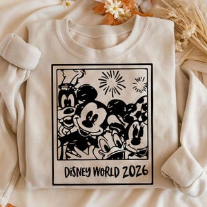 Op de afbeelding: Cr&egrave;mekleurige sweatshirt met een zwart-witte afbeelding van Disney-personages, waaronder Mickey Mouse, Minnie Mouse en Donald Duck. De afbeelding is omlijst als een foto met de tekst "Disney World 2026" onderaan.