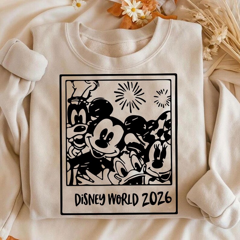 Op de afbeelding: Cr&egrave;mekleurige sweatshirt met een zwart-witte afbeelding van Disney-personages, waaronder Mickey Mouse, Minnie Mouse en Donald Duck. De afbeelding is omlijst als een foto met de tekst "Disney World 2026" onderaan.