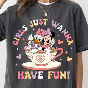 May include: A dark gray t-shirt with a graphic of a cartoon duck and mouse sitting in a teacup. The text "Girls Just Wanna Have Fun!" is printed in colorful letters on the shirt.