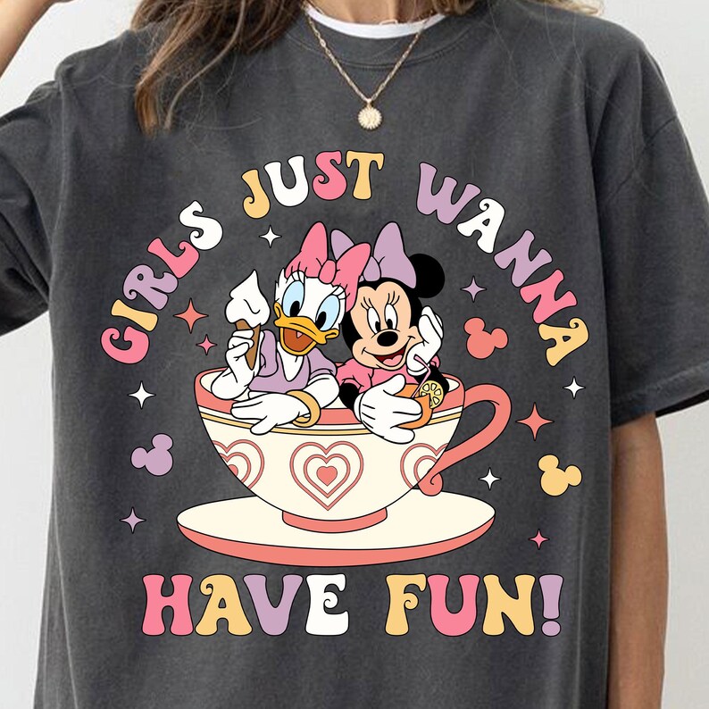 May include: A dark gray t-shirt with a graphic of a cartoon duck and mouse sitting in a teacup. The text "Girls Just Wanna Have Fun!" is printed in colorful letters on the shirt.