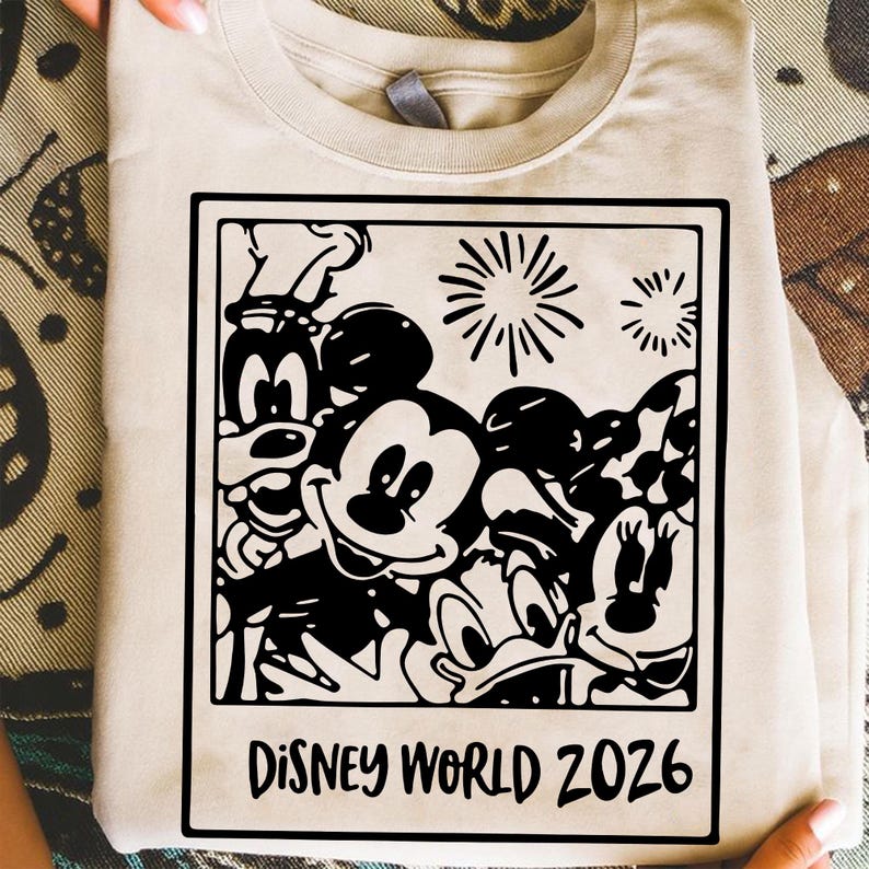 Op de afbeelding: Een beige t-shirt met een zwart-witte afbeelding van Mickey Mouse, Goofy, Donald Duck en andere Disney-personages. De afbeelding is omlijst met een zwarte rand en bevat de tekst "DISNEY WORLD 2026" aan de onderkant.