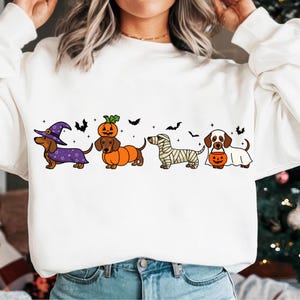 Peut inclure: Sweat-shirt blanc avec un motif de teckel sur le thème d'Halloween. L'illustration comprend des teckels déguisés en sorcière, citrouille, momie et fantôme, avec des chauves-souris et des étoiles. Un design amusant et festif pour la saison effrayante.