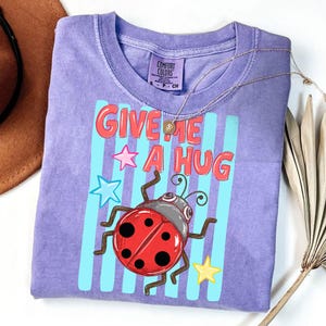 Pu&ograve; includere: Una maglietta Comfort Colors viola con la scritta "GIVE ME A HUG" in lettere rosse. La maglietta presenta una coccinella dei cartoni animati con un guscio rosso e una testa grigia, insieme a stelle blu e gialle. Una collana d'oro &egrave; drappeggiata sulla maglietta.