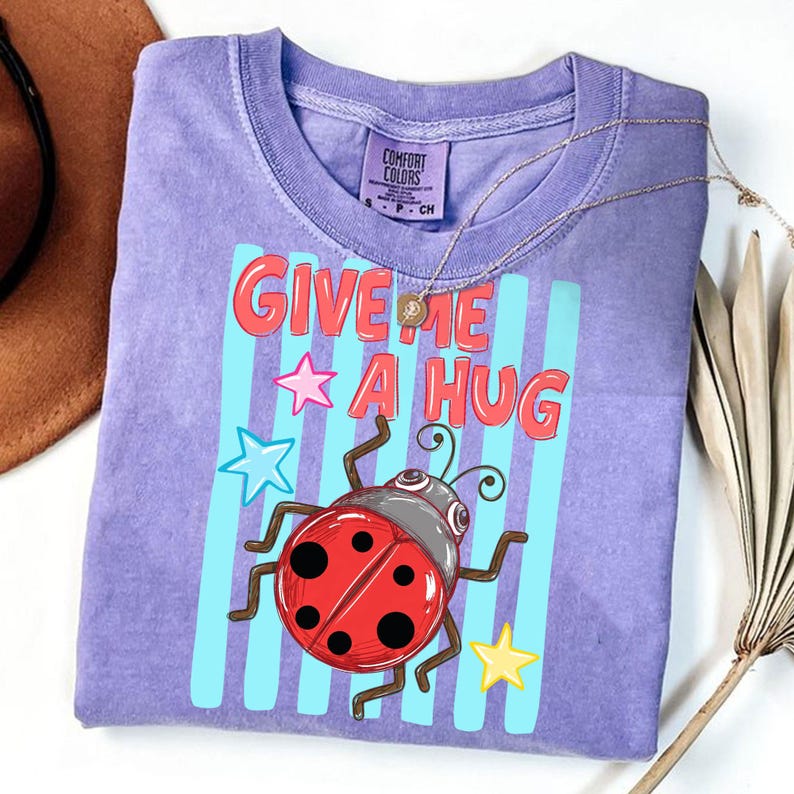 Pu&ograve; includere: Una maglietta Comfort Colors viola con la scritta "GIVE ME A HUG" in lettere rosse. La maglietta presenta una coccinella dei cartoni animati con un guscio rosso e una testa grigia, insieme a stelle blu e gialle. Una collana d'oro &egrave; drappeggiata sulla maglietta.