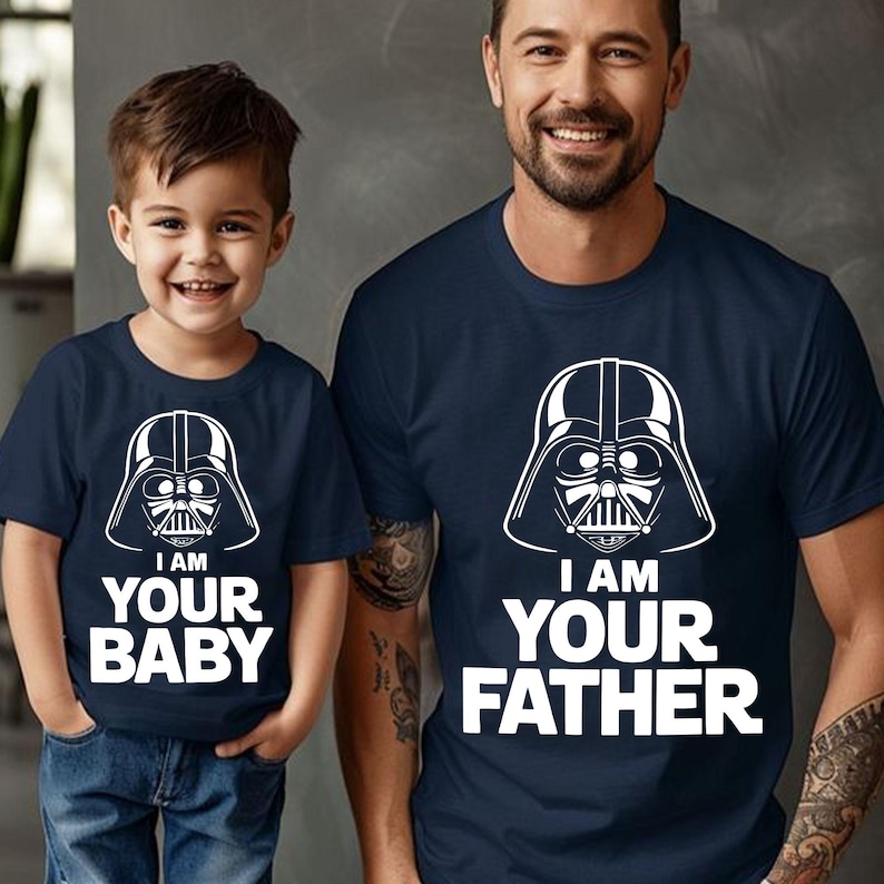 May include: Two navy blue t-shirts with white graphic of Darth Vader's helmet. One shirt says "I AM YOUR BABY", the other says "I AM YOUR FATHER". The shirts are worn by a child and an adult.