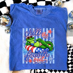 May include: A blue Comfort Colors t-shirt featuring a graphic of an alligator wearing sunglasses and a bikini, lounging on a red, white, and blue inner tube. The text "Chill the Fourth Out" is written in a script font.
