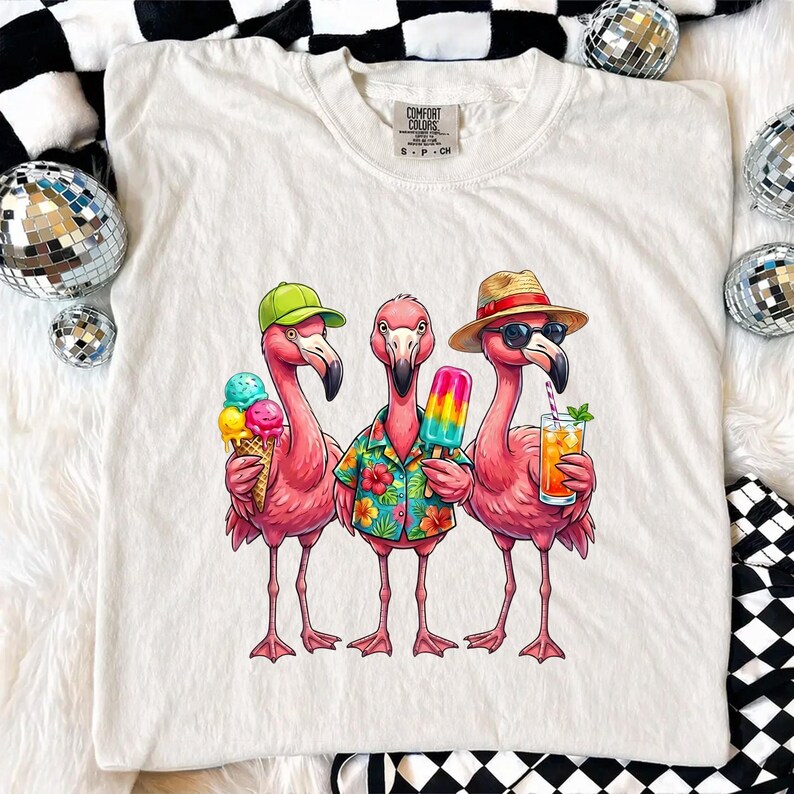 May include: White Comfort Colors t-shirt featuring a graphic of three pink flamingos. One flamingo wears a green cap and holds an ice cream cone, another wears a Hawaiian shirt and holds a popsicle, and the third wears a straw hat and sunglasses, holding a drink.