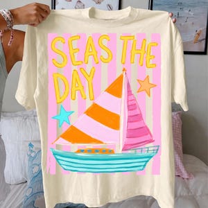 May include: Cream-colored t-shirt with a colorful sailboat graphic. The design features a sailboat with orange and pink sails, a turquoise hull, and the text "SEAS THE DAY" in yellow. Pink and white stripes form the background, with blue and orange stars.