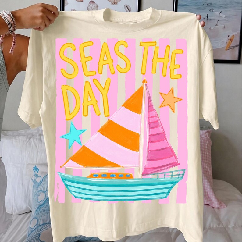 May include: Cream-colored t-shirt with a colorful sailboat graphic. The design features a sailboat with orange and pink sails, a turquoise hull, and the text "SEAS THE DAY" in yellow. Pink and white stripes form the background, with blue and orange stars.