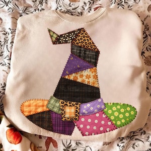 May include: Cream-coloured sweatshirt with a patchwork witch's hat design. The hat features various fabric patterns, including leopard print, plaid, and floral, in shades of orange, purple, green, and pink. The hat is stitched onto the sweatshirt.