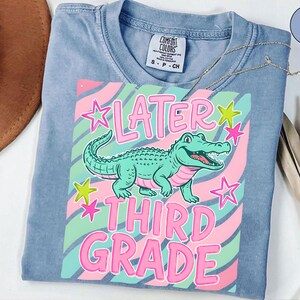 May include: A light blue t-shirt with a graphic featuring a cartoon alligator and the words "Later Third Grade" in pink. The design includes pink and green stars and wavy stripes.