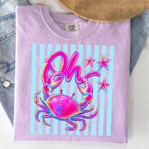 May include: A light purple t-shirt with a colorful crab graphic. The crab is pink, blue, and yellow, with the word "Oh" above it in a matching font. The design includes light blue vertical stripes and three pink stars.