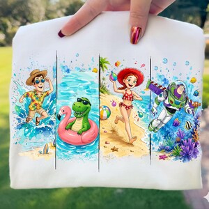 May include: White sweatshirt featuring four colorful panels with cartoon characters in a summer theme. A boy in a straw hat, a dinosaur in sunglasses, a girl in a red swimsuit, and Buzz Lightyear are depicted in beach scenes.