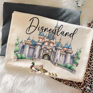 May include: A cream-colored throw pillow with the word "Disneyland" in a script font. The pillow features a detailed illustration of the Disneyland castle, ducks, and ducklings. The pillow is resting on a white, fluffy surface.