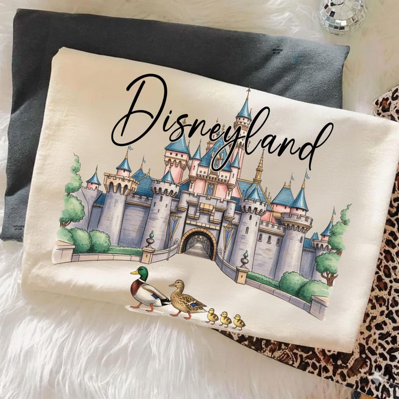 May include: A cream-colored throw pillow with the word "Disneyland" in a script font. The pillow features a detailed illustration of the Disneyland castle, ducks, and ducklings. The pillow is resting on a white, fluffy surface.