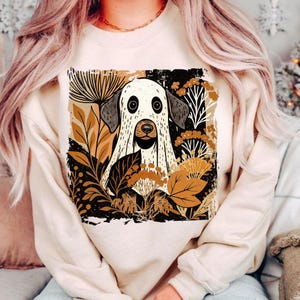 Pu&ograve; includere: Felpa beige con un'illustrazione stravagante di un cane vestito da fantasma, circondato da foglie dai toni autunnali. Il cane ha grandi occhi rotondi e un'espressione amichevole. Il disegno &egrave; su sfondo nero.