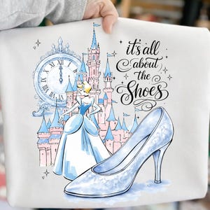 May include: A white graphic featuring Cinderella, a castle, and a glass slipper. The text "it's all about the Shoes" is written in cursive. The illustration is in shades of blue, pink, and white.