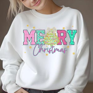 Puede incluir: Sudadera blanca con un gr&aacute;fico colorido de "MERRY Christmas". Las letras son de cuadros rosas, turquesa y rosa brillante. Un &aacute;rbol de Navidad y copos de nieve tambi&eacute;n est&aacute;n presentes en el dise&ntilde;o. Una prenda superior festiva.