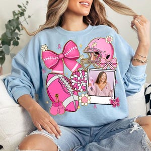 May include: Light blue sweatshirt featuring pink football-themed graphics, including a football, helmet, bow, and boxing gloves. A photo of a woman holding a pink ribbon is also included. The design incorporates pink ribbons and flowers.