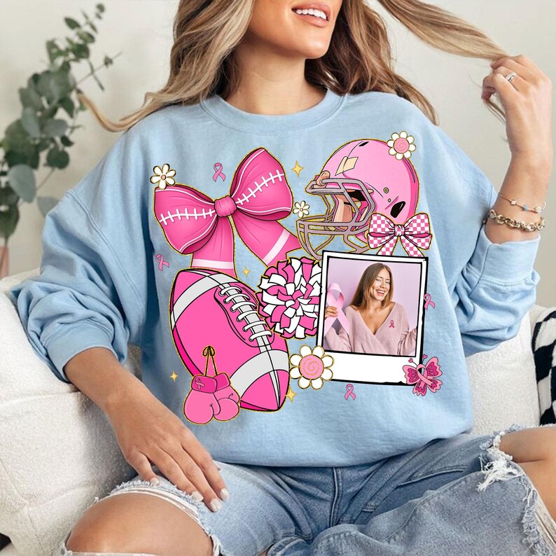 May include: Light blue sweatshirt featuring pink football-themed graphics, including a football, helmet, bow, and boxing gloves. A photo of a woman holding a pink ribbon is also included. The design incorporates pink ribbons and flowers.