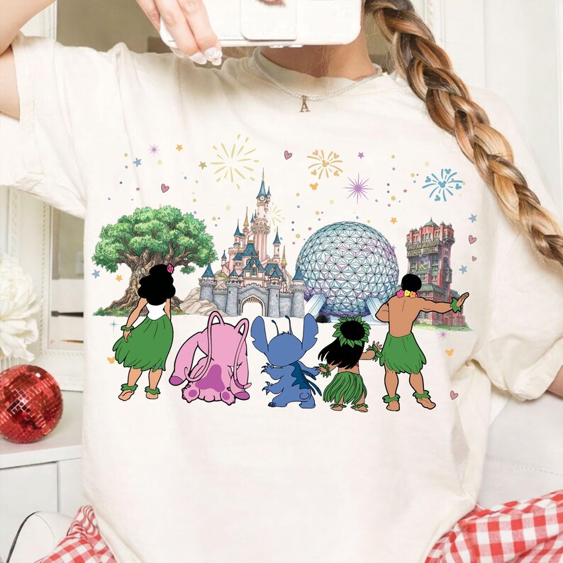 May include: Cream-colored t-shirt featuring a colorful graphic of Disney characters and landmarks. The design includes a castle, a large tree, a geodesic sphere, and cartoon figures in front of fireworks and hearts. The shirt is a casual style.