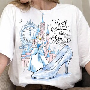 May include: White t-shirt featuring a Cinderella-themed design. The graphic includes a light blue high-heeled shoe, a castle, a clock, and the text "it's all about the Shoes". The design is in a watercolor style.