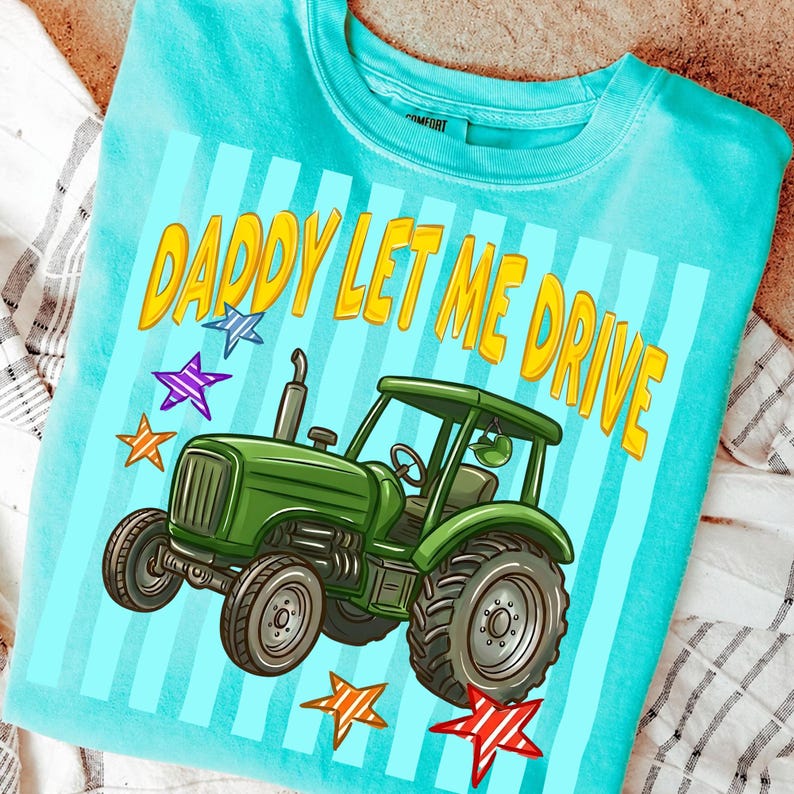 Pu&ograve; includere: T-shirt azzurra con la scritta gialla "DADDY LET ME DRIVE". Sotto il testo c'&egrave; una grafica di un trattore verde, con accenti di stelle colorate. La maglietta ha un girocollo e maniche corte.