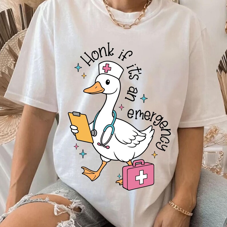 Puede incluir: Camiseta blanca con un ganso de dibujos animados que lleva un gorro de enfermera y sostiene un portapapeles y un estetoscopio. El texto "Honk if it's an emergency" est&aacute; impreso en la camiseta.