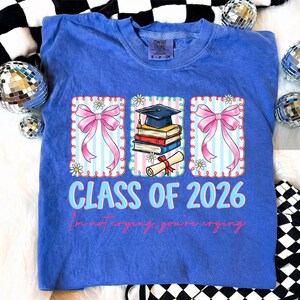 May include: A blue Comfort Colors t-shirt with a graduation-themed graphic. The graphic includes pink bows, a graduation cap, books, and a diploma. The text reads "CLASS OF 2026" and "I'm not crying, you're crying."