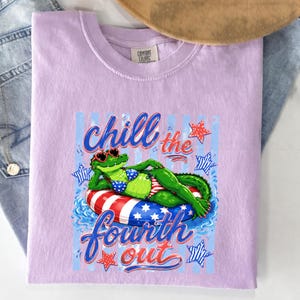 May include: Lilac Comfort Colors t-shirt featuring a cartoon alligator wearing sunglasses and a bikini, lounging on a red, white, and blue inflatable ring. The shirt has the text "Chill the Fourth Out" in a patriotic font.