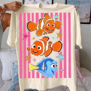 May include: Cream-colored t-shirt featuring a graphic of two orange and white clownfish and a blue and yellow fish, set against a pink striped background with star accents. The design is a cartoon style.