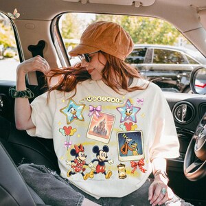May include: Cream-colored sweatshirt featuring Disney characters. The design includes images of Mickey and Minnie Mouse, Donald and Daisy Duck, Goofy, and a castle. The word "Magical" is arched across the top. The sweatshirt has a relaxed fit.