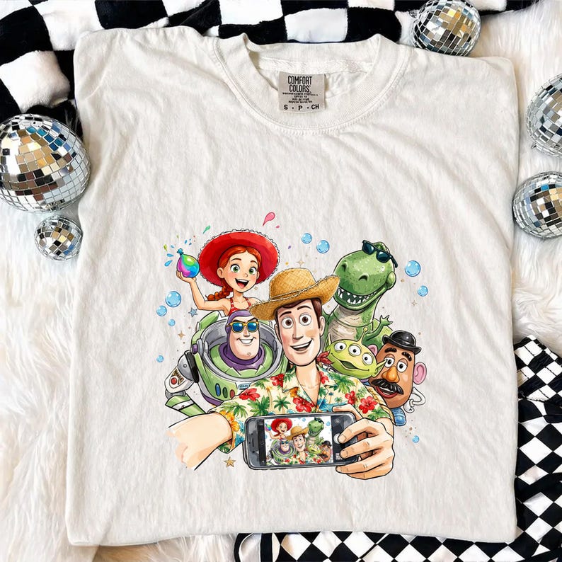 May include: Cream-colored t-shirt featuring a colorful graphic of Toy Story characters taking a selfie. The design includes Woody, Buzz Lightyear, Jessie, Rex, Mr. Potato Head, and the aliens. The shirt has a classic crew neck and short sleeves.