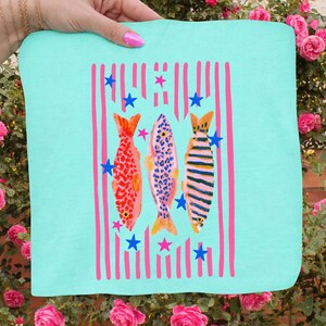 Pode incluir: Um tecido quadrado azul claro com um design de peixes coloridos. Tr&ecirc;s peixes s&atilde;o retratados com padr&otilde;es vermelhos, azuis e listrados, em fundo de listras verticais rosa e estrelas azuis. O tecido &eacute; segurado por uma m&atilde;o.
