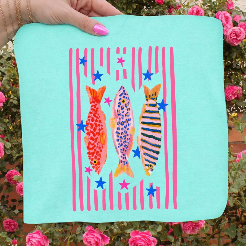 Pode incluir: Um tecido quadrado azul claro com um design de peixes coloridos. Tr&ecirc;s peixes s&atilde;o retratados com padr&otilde;es vermelhos, azuis e listrados, em fundo de listras verticais rosa e estrelas azuis. O tecido &eacute; segurado por uma m&atilde;o.