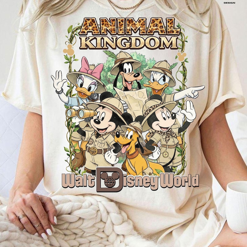 May include: Cream-colored t-shirt featuring a graphic of Disney characters in safari attire. The design includes Mickey Mouse, Minnie Mouse, Donald Duck, Daisy Duck, Goofy, and Pluto, with the text "ANIMAL KINGDOM" and "Walt Disney World".